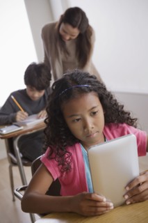 Schoolgirl (10-11) holding digital tablet with boy (12-13) and teacher in background. 
Photo : Rob