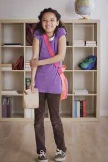 Full length of schoolgirl (10-11) standing in classroom holding paper bag. 
Photo : Rob Lewine