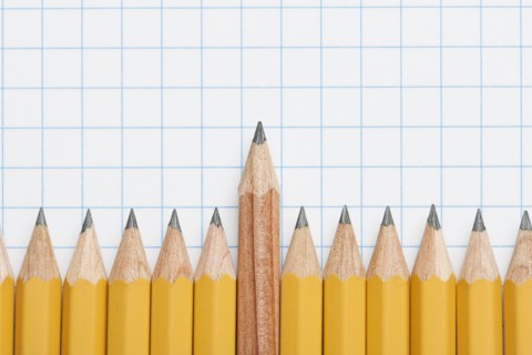 Single wooden pencil rising above row of sharpened pencils on graph paper, studio shot. Photo:
