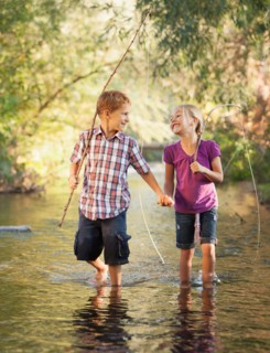 Little boy (6-7) and little girl (4-5) holding hands and walking together in small stream holding wooden stick fishing poles