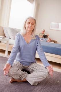 Senior woman practicing yoga with husband reading in background. Photo : Rob Lewine