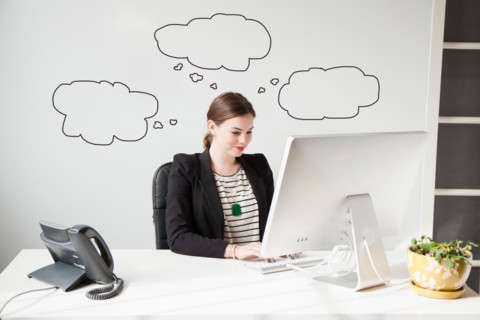 Studio shot of young woman working at desk with thought bubbles around her head. Photo : Jessica