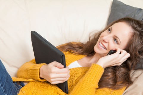 Woman lying on sofa with digital tablet and talking via cell phone