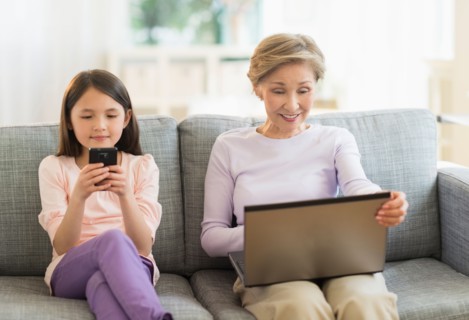 Granddaughter (8-9) and grandmother sitting on sofa and using laptop and cell phone.