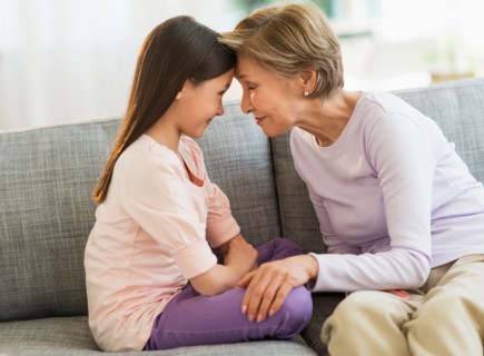 Granddaughter (8-9) and grandmother sitting on sofa and touching with heads.