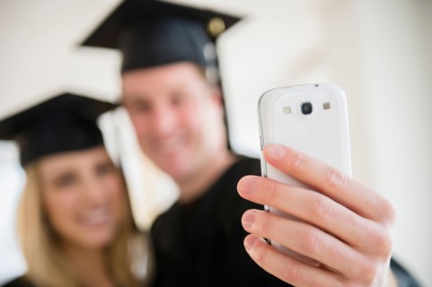 Couple of friends in graduation gowns taking photo with smartphone