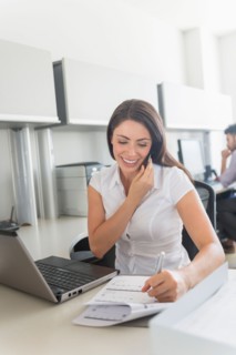 Woman talking on cell phone and doing paperwork in office, man in background.