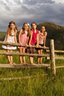 Four girlfriends (6-7, 8-9, 10-11) standing on wooden fence in grass