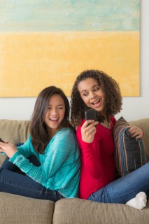Two friends (12-13,16-17) sitting on sofa and playing with smartphone