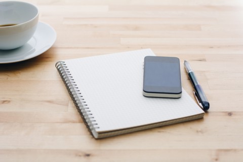 Spiral notebook, Ballpoint pen, Mobile phone and coffee cup on wooden table.
Photo : Kristin Lee