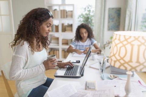 Woman using laptop at home, girl (8-9) doing homework in background.
