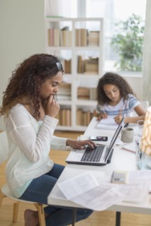 Woman using laptop at home, girl (8-9) doing homework in background.