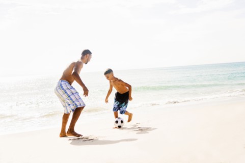 Father and son (10-11) playing soccer on beach