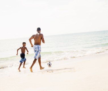 Father and son (10-11) playing soccer on beach