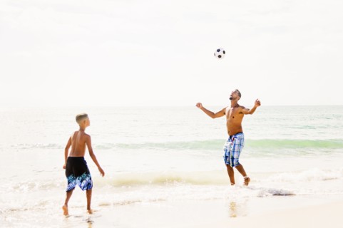 Father and son (10-11) playing soccer on beach