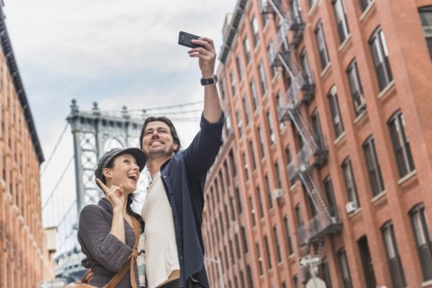 Couple taking selfie on street, Brooklyn Bridge in background. Brooklyn, New York.