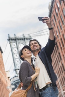 Couple taking selfie on street, Brooklyn Bridge in background. Brooklyn, New York.