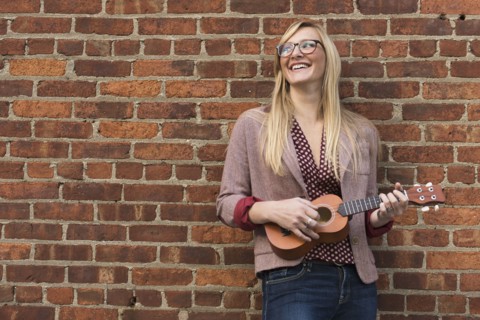 Woman standing against brick wall and playing ukulele.