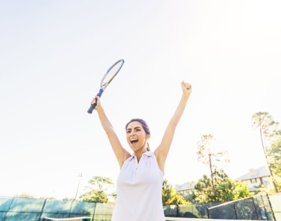 Portrait of young woman holding tennis racket, raising arms in celebration