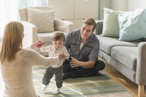 Happy parents helping little son (2-3 years) walking in living room.