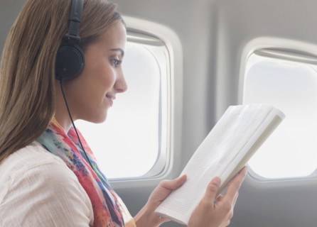Young woman wearing headphones while reading book on plane.