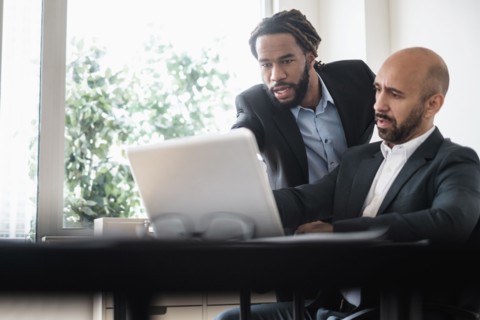 Two businessmen working with laptop by desk in office.