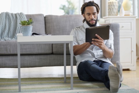 Young man with dreadlocks using digital table by sofa in living room.