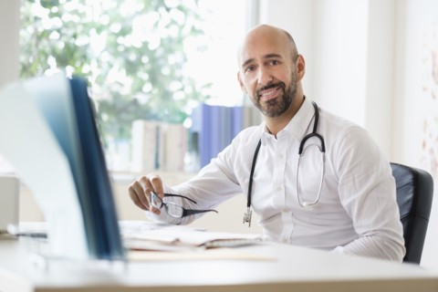 Portrait of smiley doctor working with laptop at desk in office.