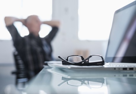 Eyeglasses and laptop on desk in office, man relaxing in background.
