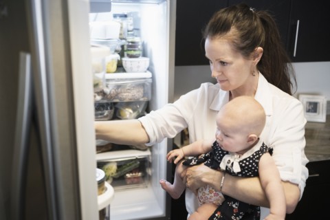 Mother looking for food in refrigerator while holding baby daughter (2-5 months).