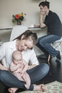 Mother holding baby (2-5 months) and father working in background.