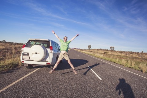 Australia, Outback, Northern Territory, Red Centre, Uluru-Kata Tjuta National Park, Woman jumping next to SUV car on road in wilderness