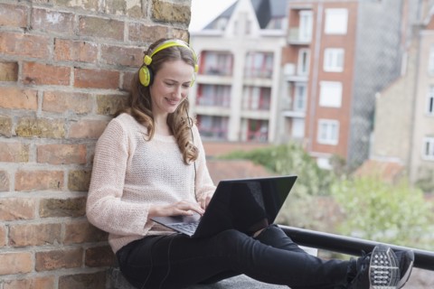 Mid-adult woman with headphones on sitting on balcony railing and using laptop