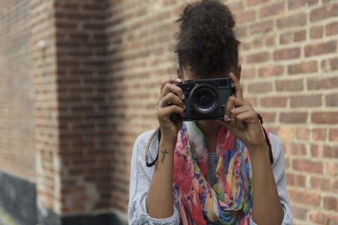Young woman taking picture with digital camera against brick wall.