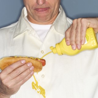 Mature man holding hotdog and accidentally spilling mustard on shirt.