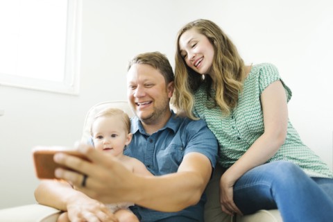 Mother and father with baby girl (12-17 months) sitting in living room and taking selfie