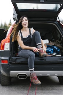 Smiling woman sitting in car trunk and tying hiking shoes