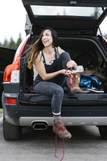 Smiling woman sitting in car trunk and tying hiking shoes