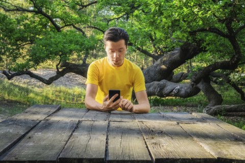 Ukraine, Dnepropetrovsk region, Novomoskovsk district, Man sitting at wooden table and using smart phone with Oak (Quercus) tree in background
