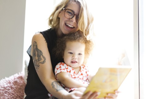 Mother sitting with daughter (12-17 months) on windowsill and reading book