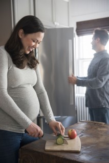 Pregnant woman cutting cucumber, man looking into refrigerator