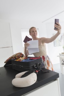 Happy mature woman packing suitcase for vacations and taking selfie