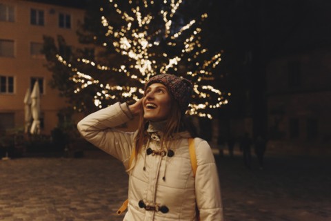 Smiling woman wearing coat by fairy lights in tree at night