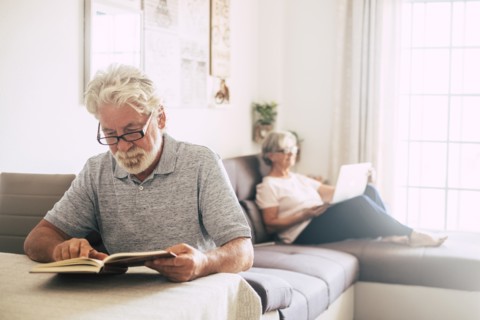 Senior man reading book as his wife uses laptop in living room