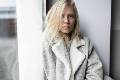 young woman in wool coat leaning against wall by window