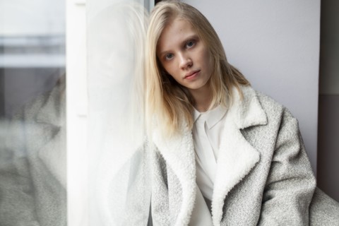 young woman in wool coat leaning against wall by window