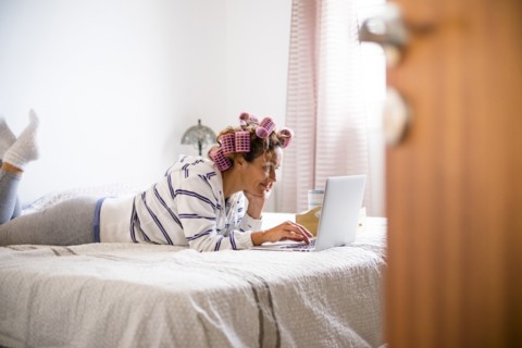 Woman with hair curlers lying on bed and using laptop