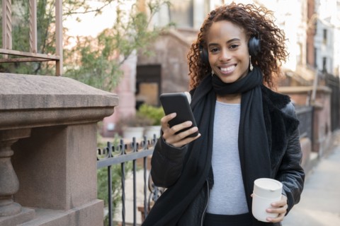 Smiling woman wearing headphones holding smart phone and cup