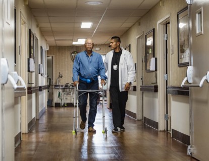 Male nurse assisting senior man walking down corridor with mobility walker