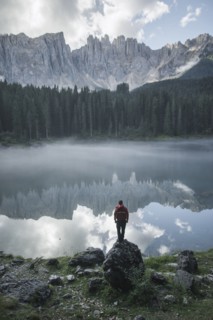 Italy, Carezza, Young man standing on rock at Lago di Carezza in Dolomite Alps at dawn